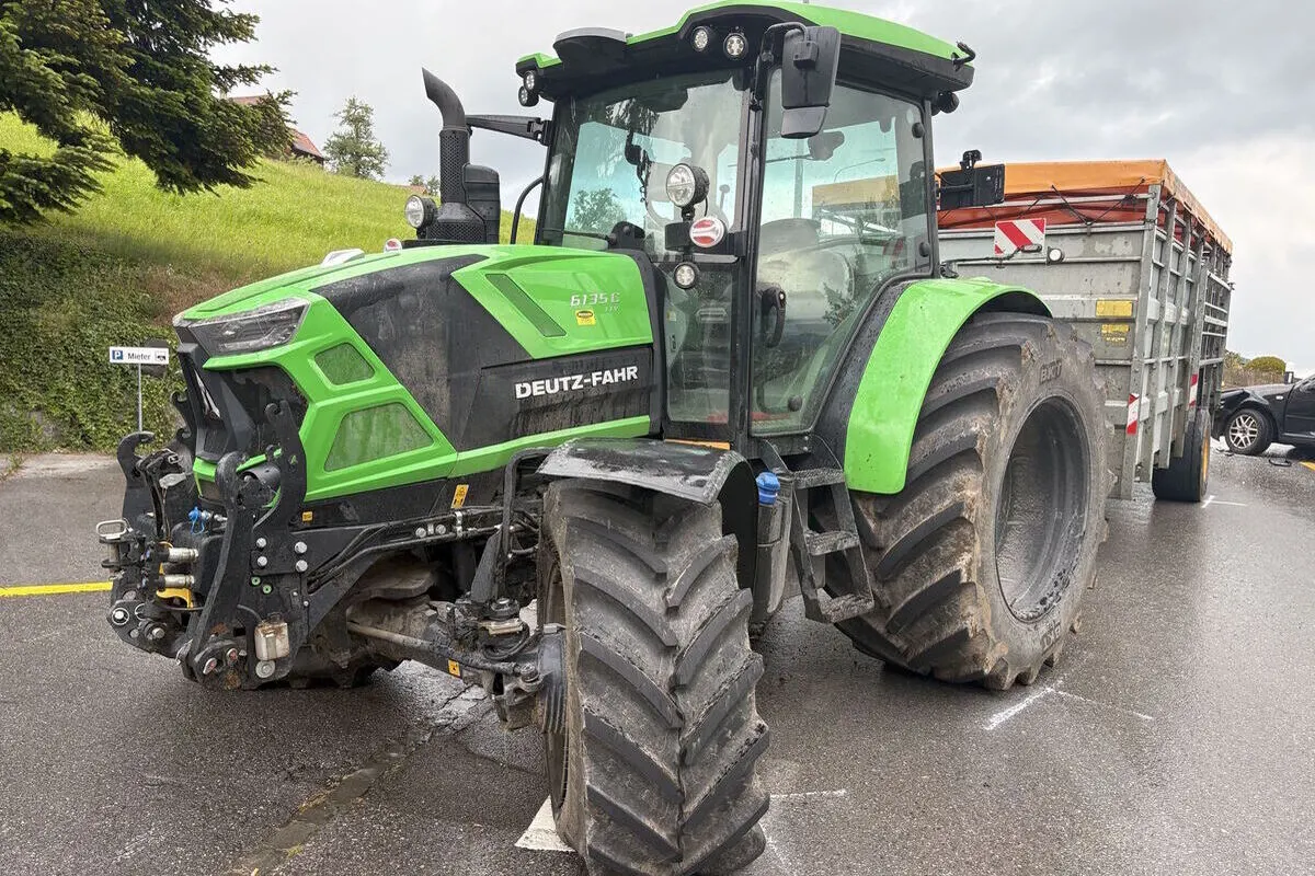 Ein grüner Traktor der Marke Deutz-Fahr steht quer über eine Straße und ist mit einem Anhänger verbunden. Im Hintergrund sind Bäume und eine grasbewachsene Böschung zu sehen.