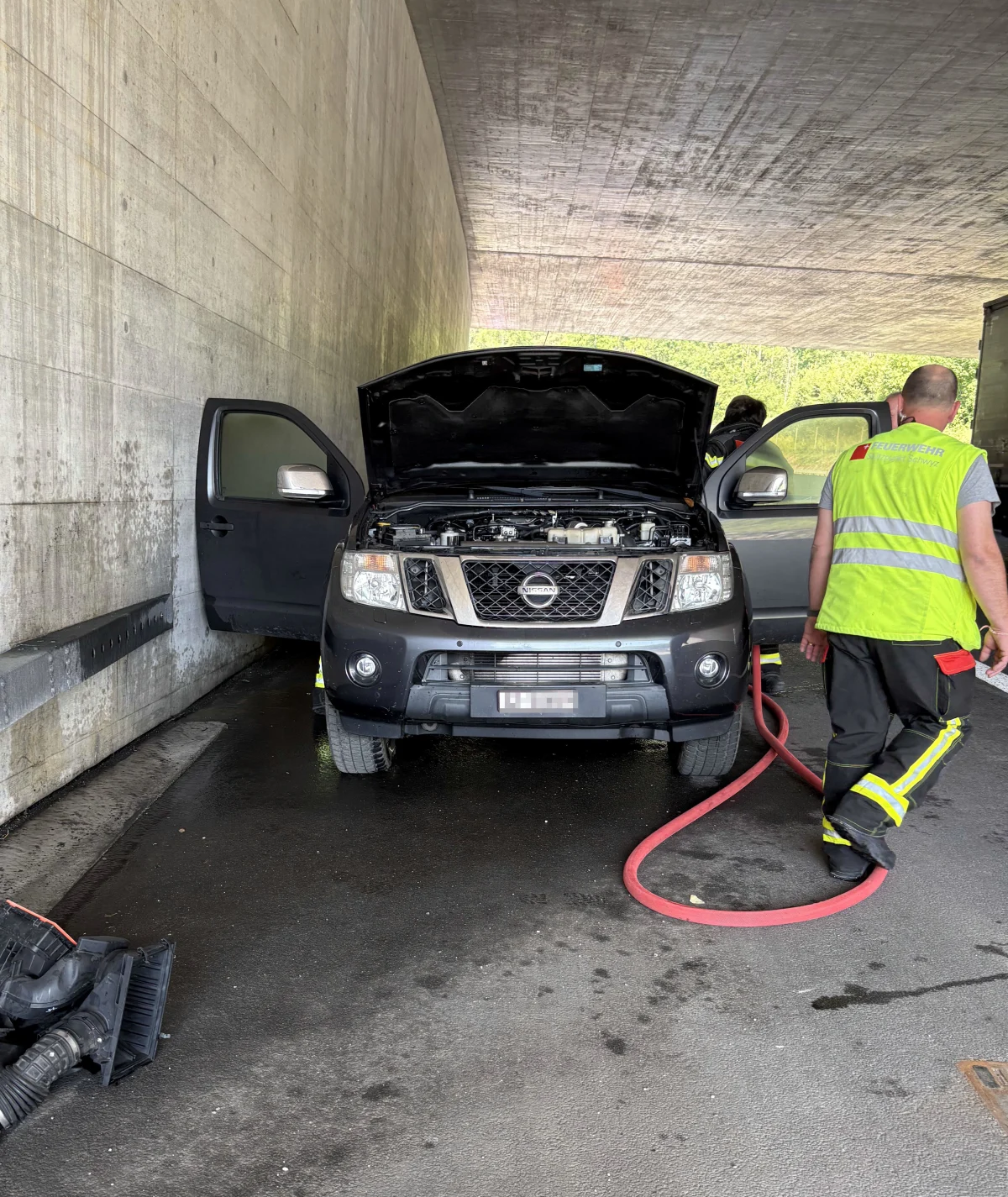 Ein schwarzer Nissan mit geöffnetem Motorraum steht unter einer Brücke. Ein Feuerwehrmann in Warnweste steht daneben.