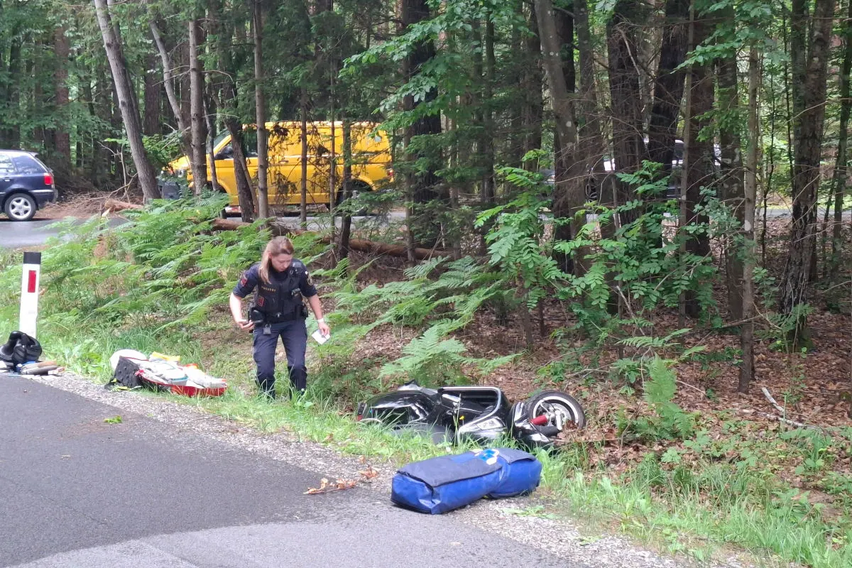 Ein Polizist untersucht ein verunfalltes Motorrad, das im Straßengraben eines Waldstücks liegt. Im Hintergrund sind ein gelber Transporter und ein weiteres Fahrzeug zu sehen.