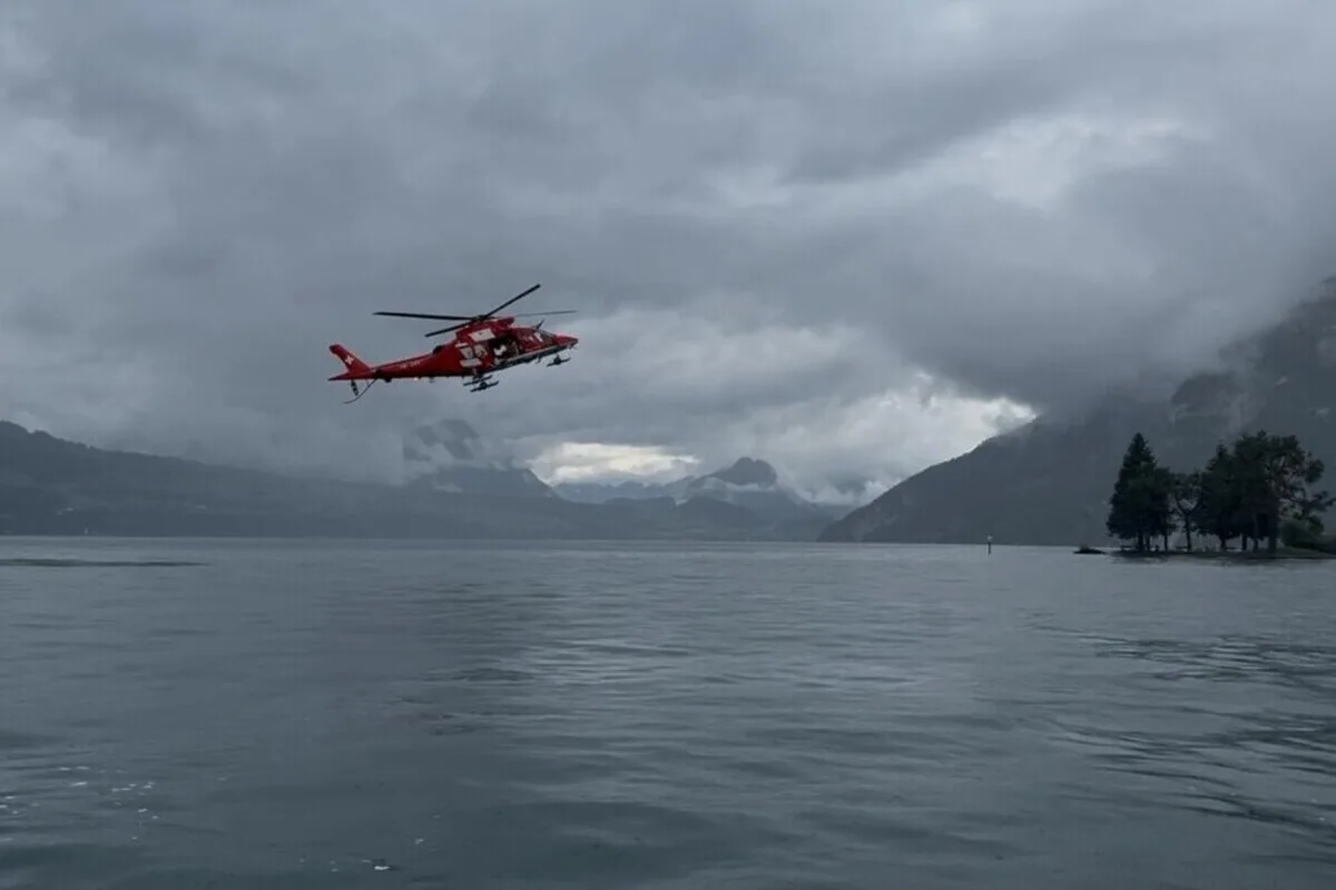 Ein roter Rettungshubschrauber schwebt über einem ruhigen See unter einem bewölkten Himmel. Im Hintergrund sind Berge sichtbar.