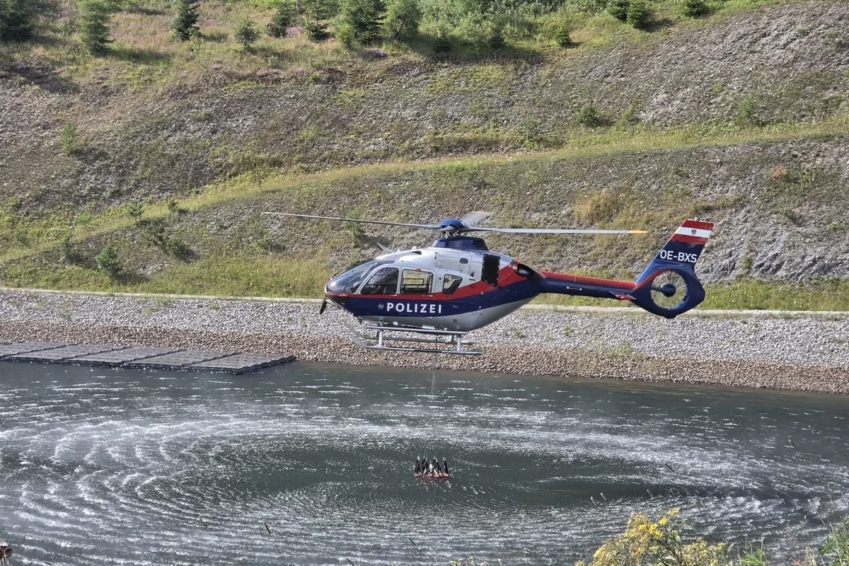 Ein Polizeihubschrauber mit der Aufschrift 'Polizei' schwebt über einem Fluss. Im Hintergrund ist ein bewachsener Hang zu sehen.
