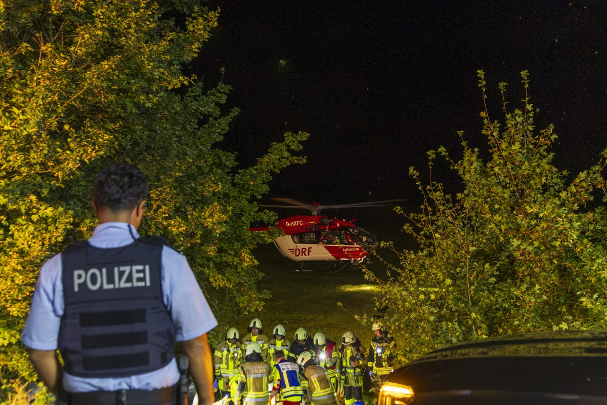 Ein Polizist und Feuerwehrleute sind nachts vor einem gelandeten Rettungshubschrauber im Einsatz. Der Hubschrauber ist im Hintergrund zu sehen.
