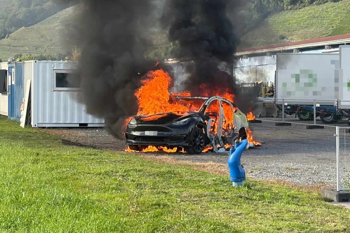 Ein schwarzes Auto brennt auf einem Schotterplatz neben Containern, wobei dichter, schwarzer Rauch aufsteigt.