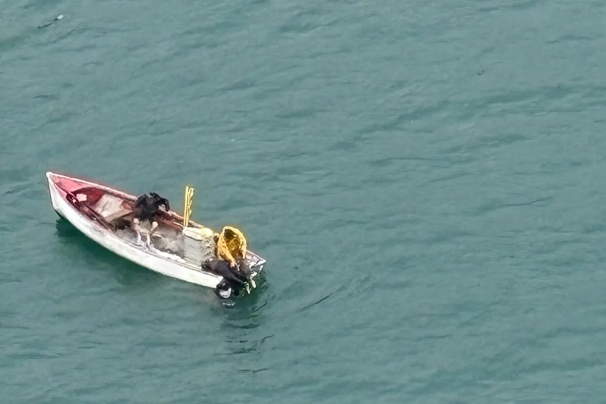 Ein kleines, baufälliges Boot befindet sich inmitten eines Gewässers. Zwei Personen und einige Gegenstände sind an Bord sichtbar.