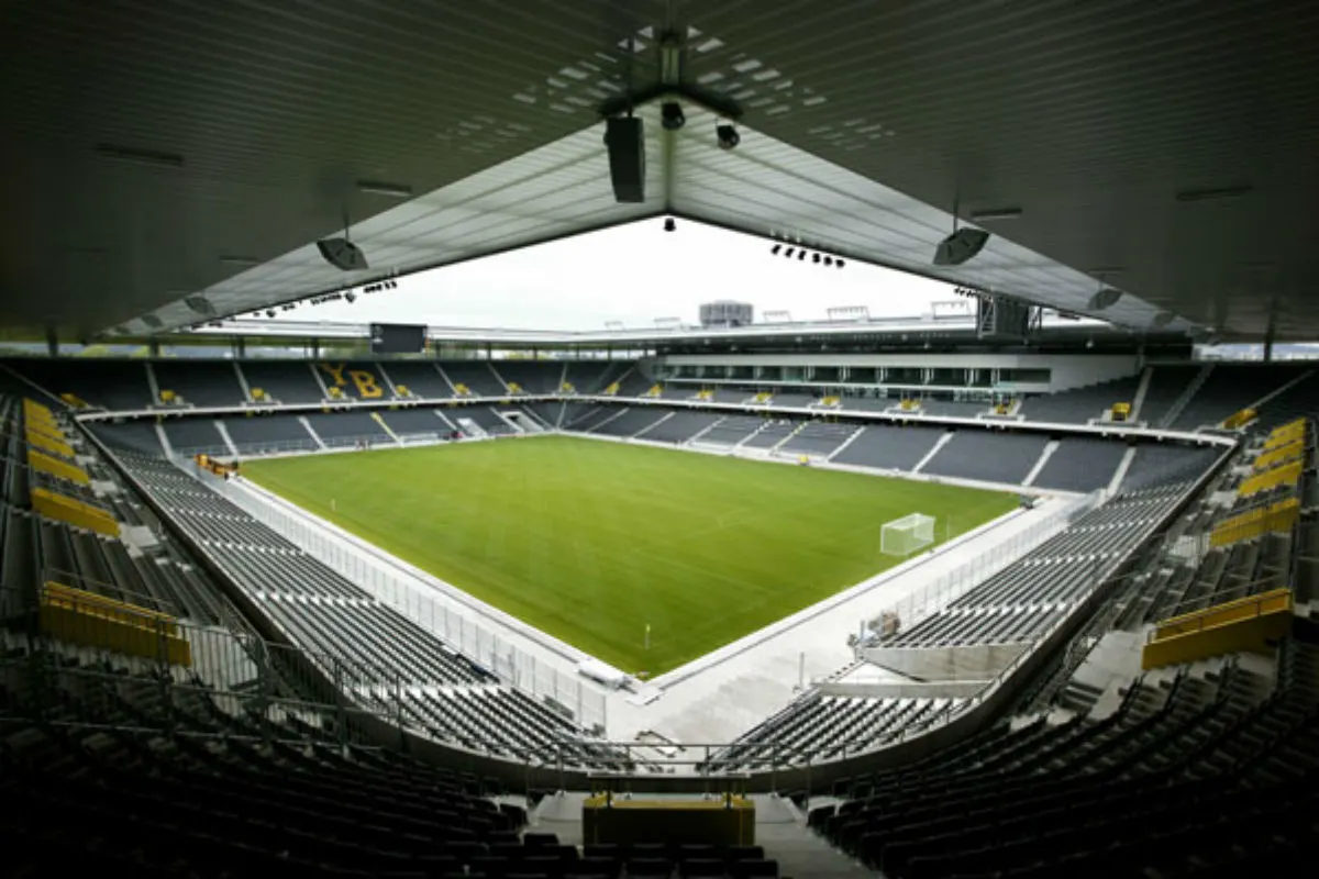 Ein leeres Fußballstadion mit grüner Rasenfläche, umgeben von grauen Sitzplätzen und einem YB-Logo an den Tribünen.