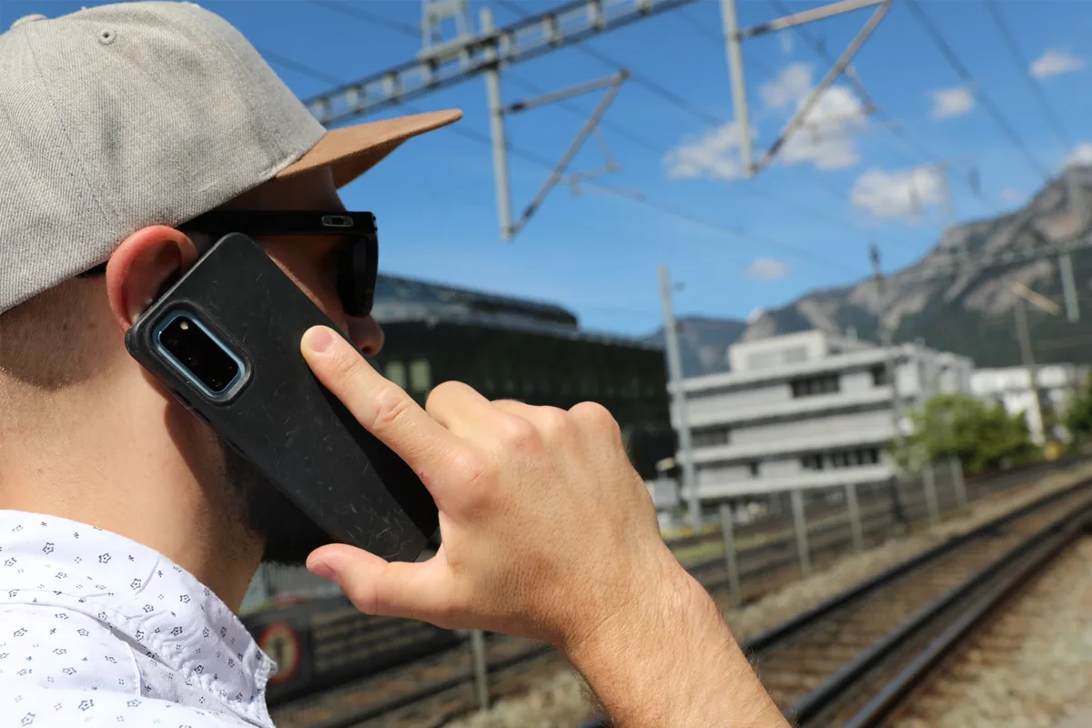 Ein Mann mit Sonnenbrille und Baseballkappe telefoniert mit einem Smartphone an einem Bahngleis. Im Hintergrund sind Gebäude und Berge zu sehen.