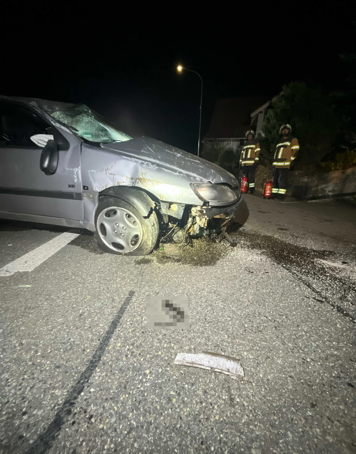 Ein graues Auto ist an der vorderen rechten Seite stark beschädigt. Im Hintergrund sind zwei Personen in Schutzkleidung zu sehen.