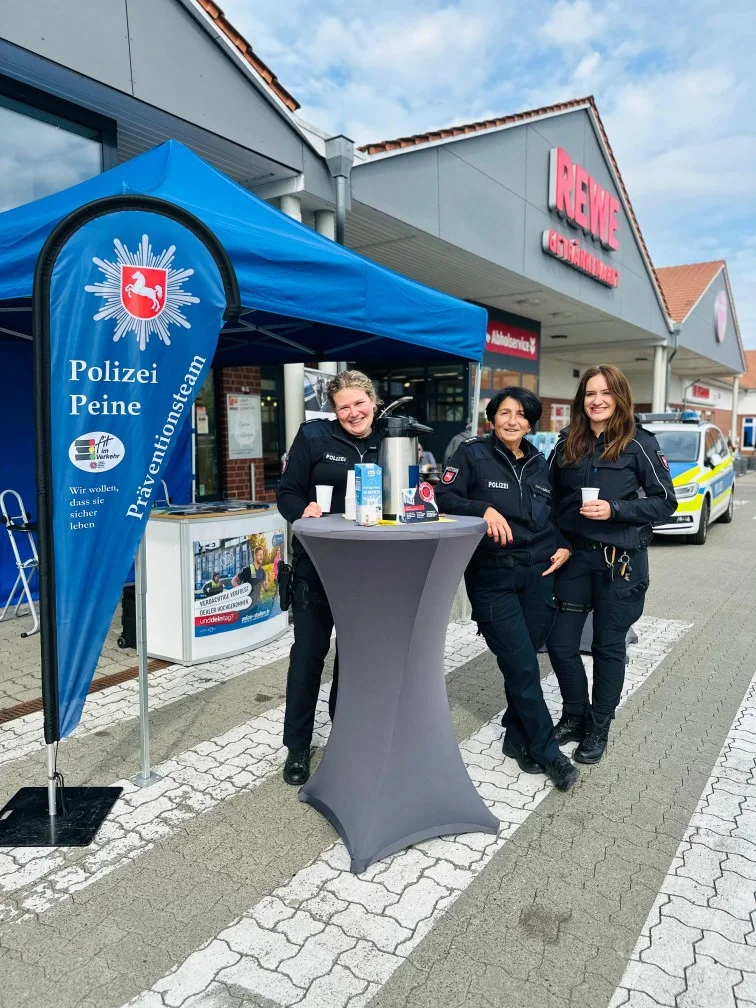 Drei Polizeibeamtinnen lächeln an einem Infostand vor einem REWE-Markt. Daneben steht ein Polizeiwagen.