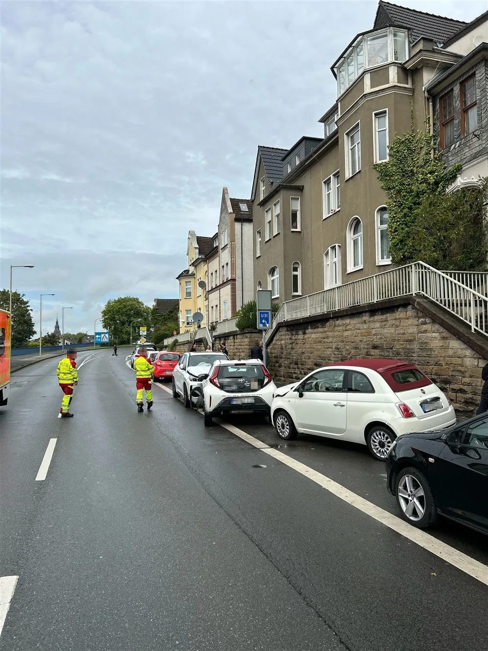 Eine Reihe geparkter Autos mit sichtbaren Schäden nach einem Auffahrunfall. Zwei Einsatzkräfte sind auf der Straße zu sehen.