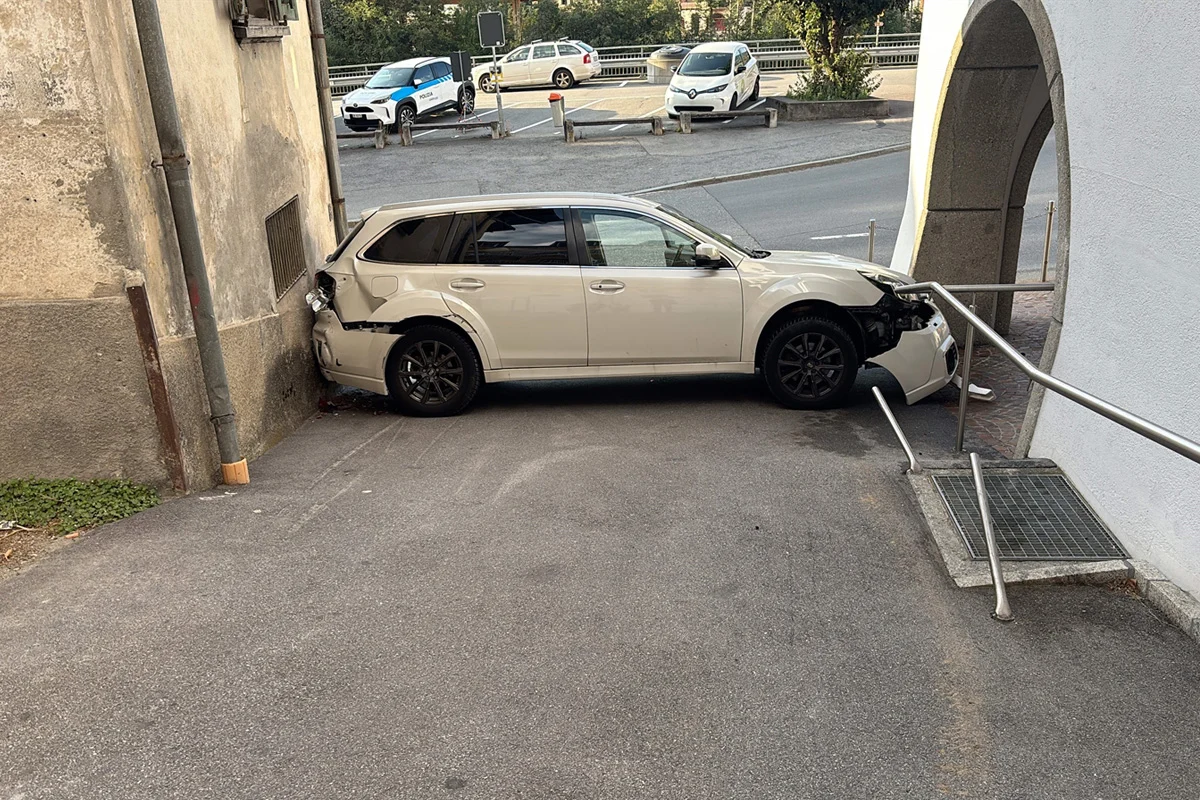 Ein weißes Auto steht mit beschädigter Front und Heck im rechten Winkel zwischen einer Mauer und einer Rampe. Im Hintergrund sind weitere Fahrzeuge sichtbar.