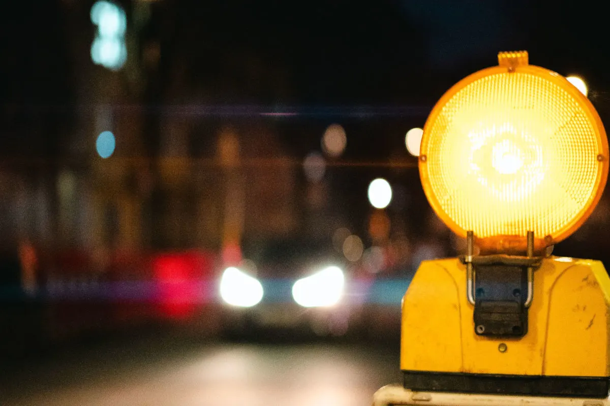 Im Vordergrund leuchtet eine gelbe Warnlampe bei Nacht, undeutliche Autoscheinwerfer sind im Hintergrund erkennbar.