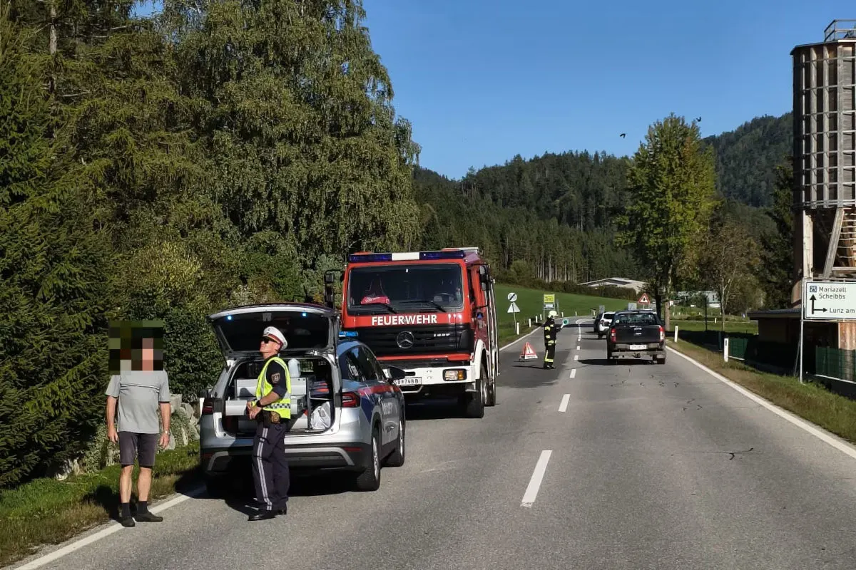 Ein Feuerwehrfahrzeug steht auf einer Landstraße, ein Polizist spricht mit einer Person neben einem Auto, bei sonnigem Wetter.