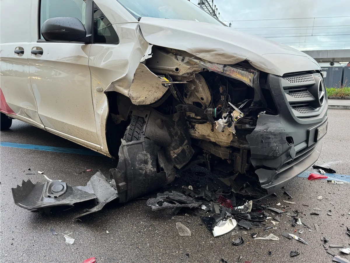 Die Vorderseite eines weißen Mercedes-Transporters ist stark beschädigt nach einer Kollision. Trümmerteile liegen auf dem Boden. Die Szene scheint an einem regnerischen Tag in einem urbanen Umfeld aufgenommen zu sein.