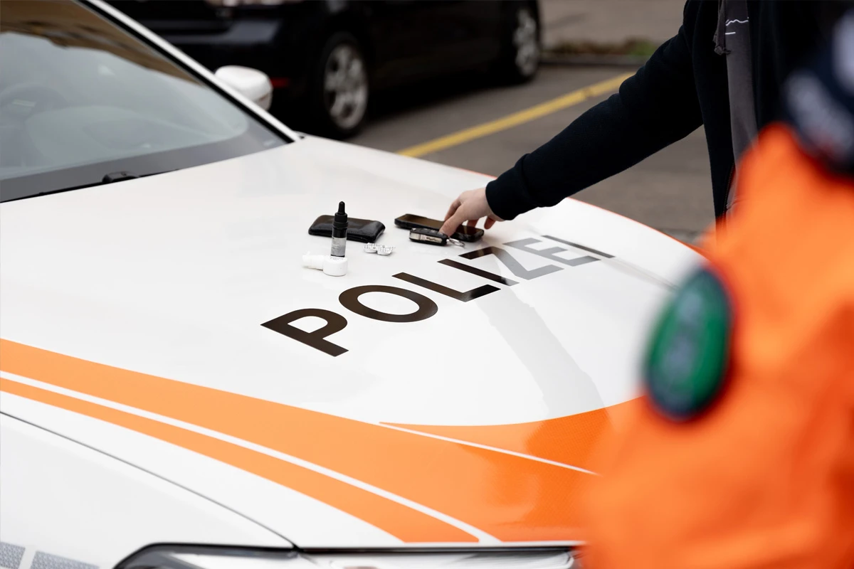 Ein weißes Polizeiauto mit dem Schriftzug 'Polizei' und orangen Streifen zeigt mehrere Gegenstände auf der Motorhaube. Ein Arm greift nach einem der Gegenstände.