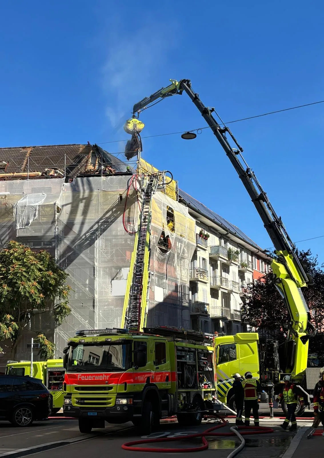 Mehrere Feuerwehrfahrzeuge sind im Einsatz, um einen Brand am Dach eines Wohnhauses in einer Stadt zu löschen. Leitern sind ausgefahren und Feuerwehrleute arbeiten vor Ort.