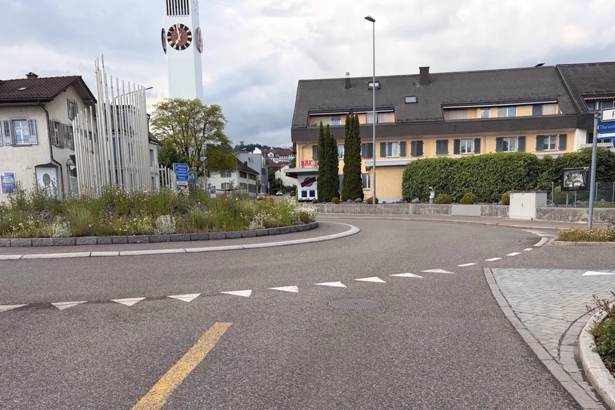 Ein leeres Kreisverkehr mit Pflanzen in der Mitte, flankiert von Gebäuden und einem großen Kirchturm im Hintergrund. Der Himmel ist bewölkt.