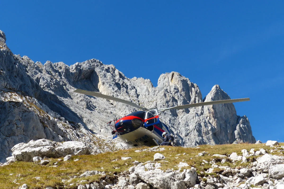 Ein blau-roter Hubschrauber ist in einer Gebirgslandschaft gelandet. Im Hintergrund sind felsige Gipfel sichtbar.