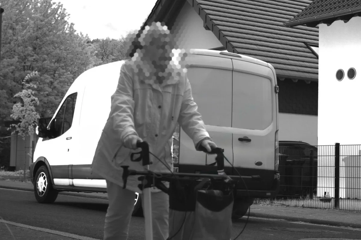 Eine ältere Person mit einem Rollator bewegt sich auf einer Straße vor einem geparkten weißen Transporter. Im Hintergrund sind Wohnhäuser und Bäume zu sehen.