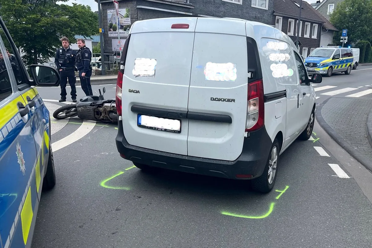 Ein weißer Dacia-Lieferwagen steht neben einem umgestürzten Motorrad auf der Straße, während zwei Polizisten in Uniform die Szenerie beobachten. Polizeifahrzeuge sind vor Ort.