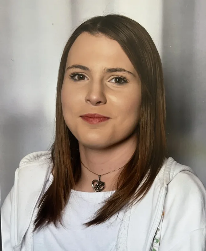 Porträt einer jungen Frau mit braunen Haaren und weißem Oberteil vor einem grauen Hintergrund. Sie trägt eine Halskette.
