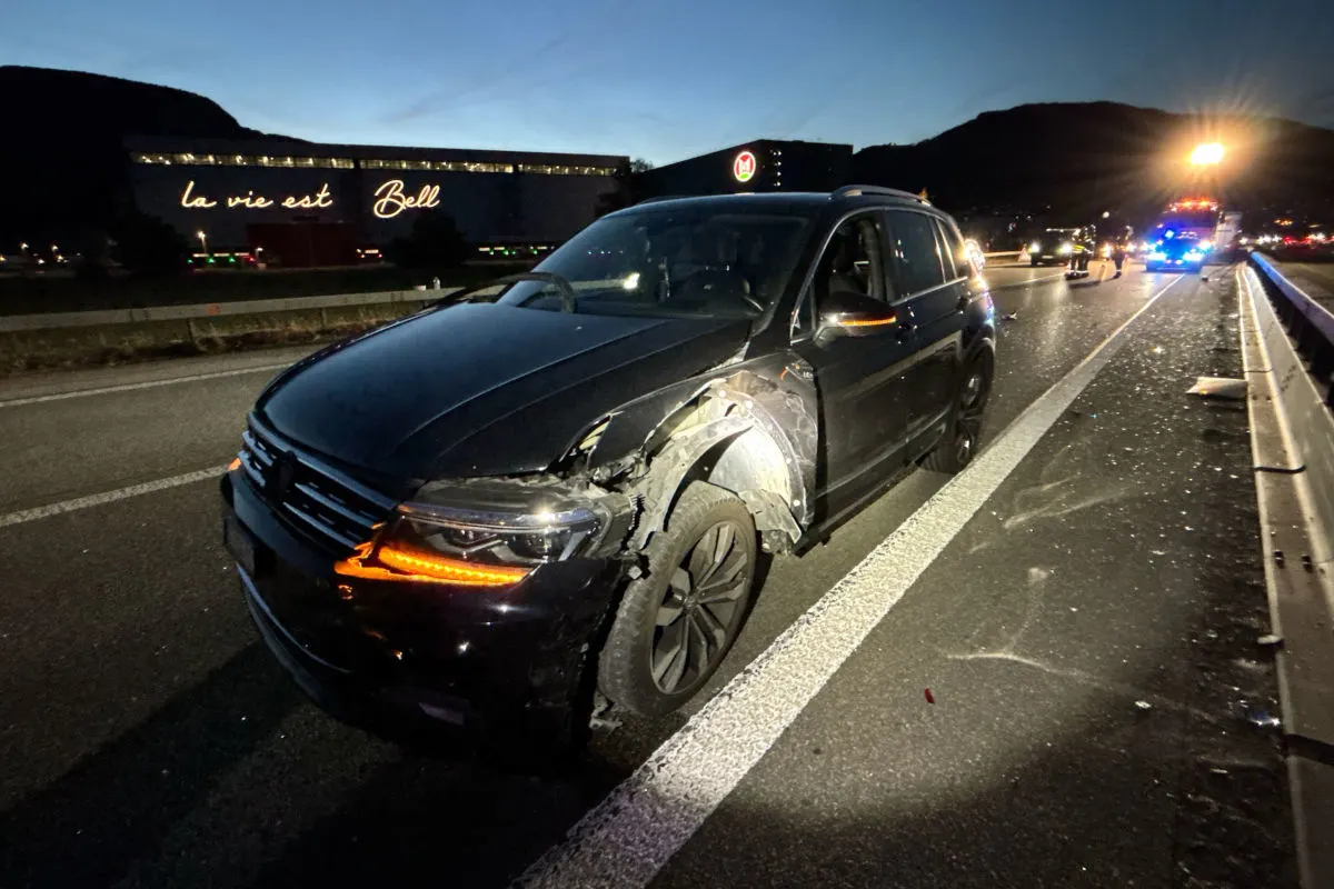 Ein dunkles Auto mit Frontschaden steht auf der Autobahn. Im Hintergrund sind Lichter eines Gebäudes erkennbar.