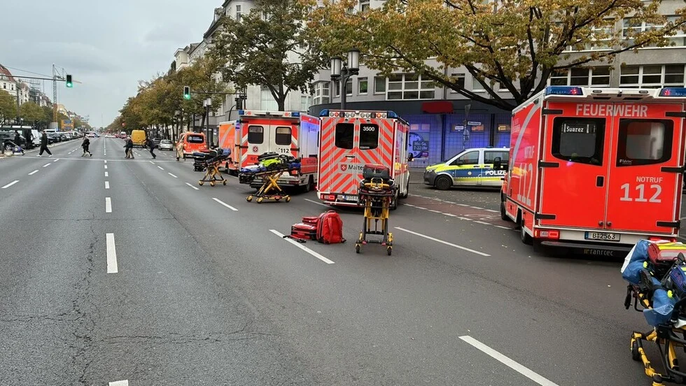 Mehrere Rettungsfahrzeuge parken auf einer Stadtstraße mit einem Polizeiauto in der Nähe. Tragen warten auf dem Bürgersteig.