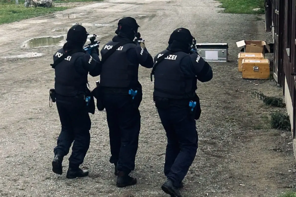 Drei Polizisten tragen Schutzkleidung und Waffen, während sie auf einem Kiesweg bei einem Einsatz vorgehen. Im Hintergrund sind Kisten an einer Gebäudewand erkennbar.