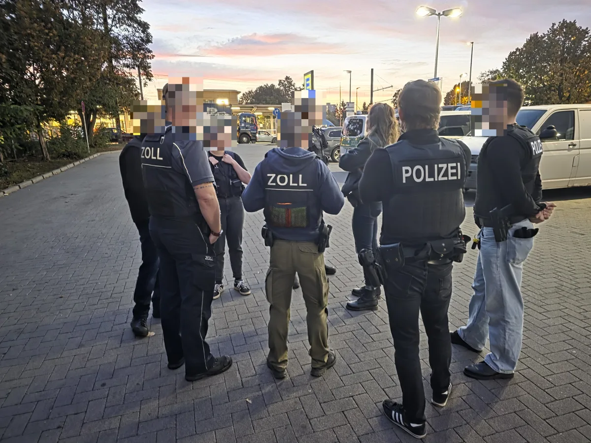 Mehrere Polizei- und Zollbeamte stehen in einer Gruppe auf einem Parkplatz und diskutieren bei einsetzender Dämmerung. Im Hintergrund sind Fahrzeuge zu sehen.