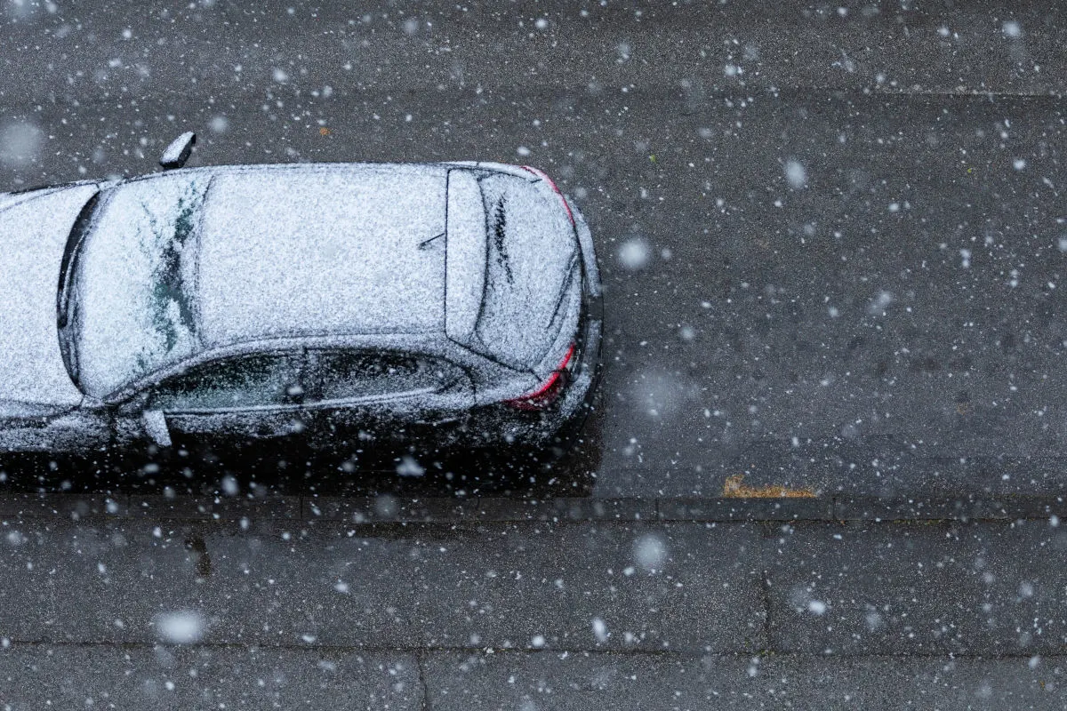 Ein Auto steht auf einer Straße und ist fast vollständig mit Schnee bedeckt. Schneeflocken fallen und die Straße wirkt nass.