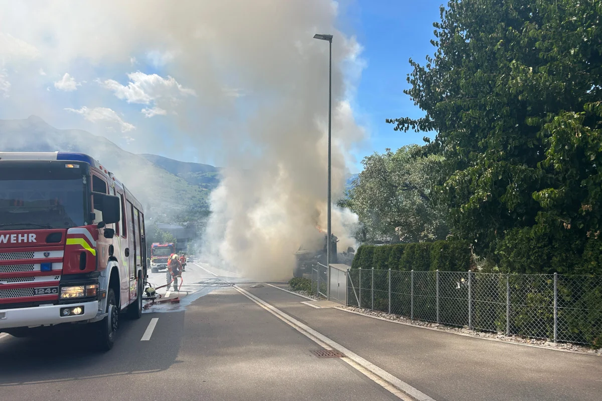 Ein großes Feuerwehrfahrzeug steht auf einer Straße, dahinter steigt dichter Rauch auf und Feuerwehrleute sind im Einsatz.