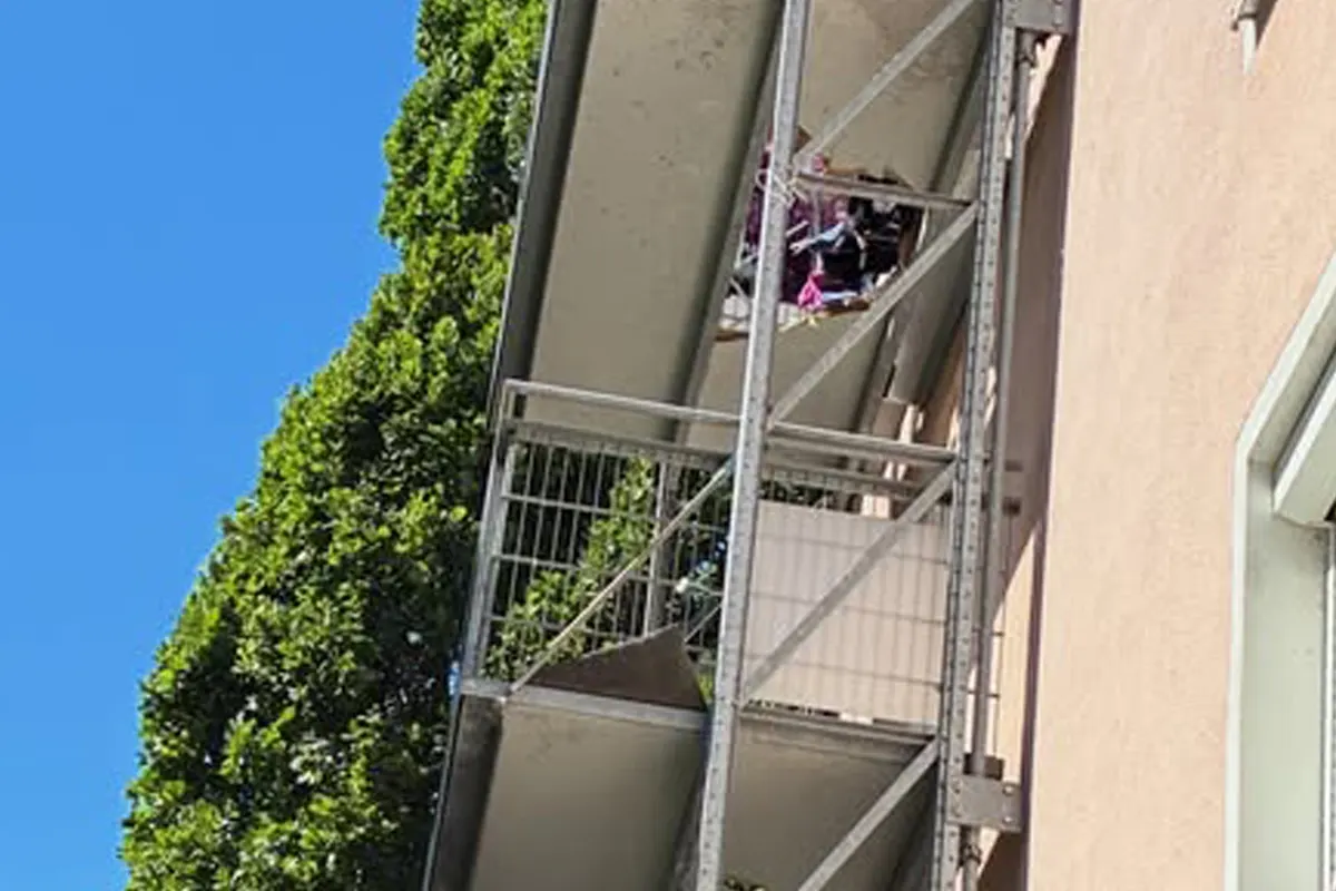 Ein beschädigter Balkon an einem Mehrfamilienhaus ist zu sehen. Der Himmel ist blau und ein Baum ist sichtbar.