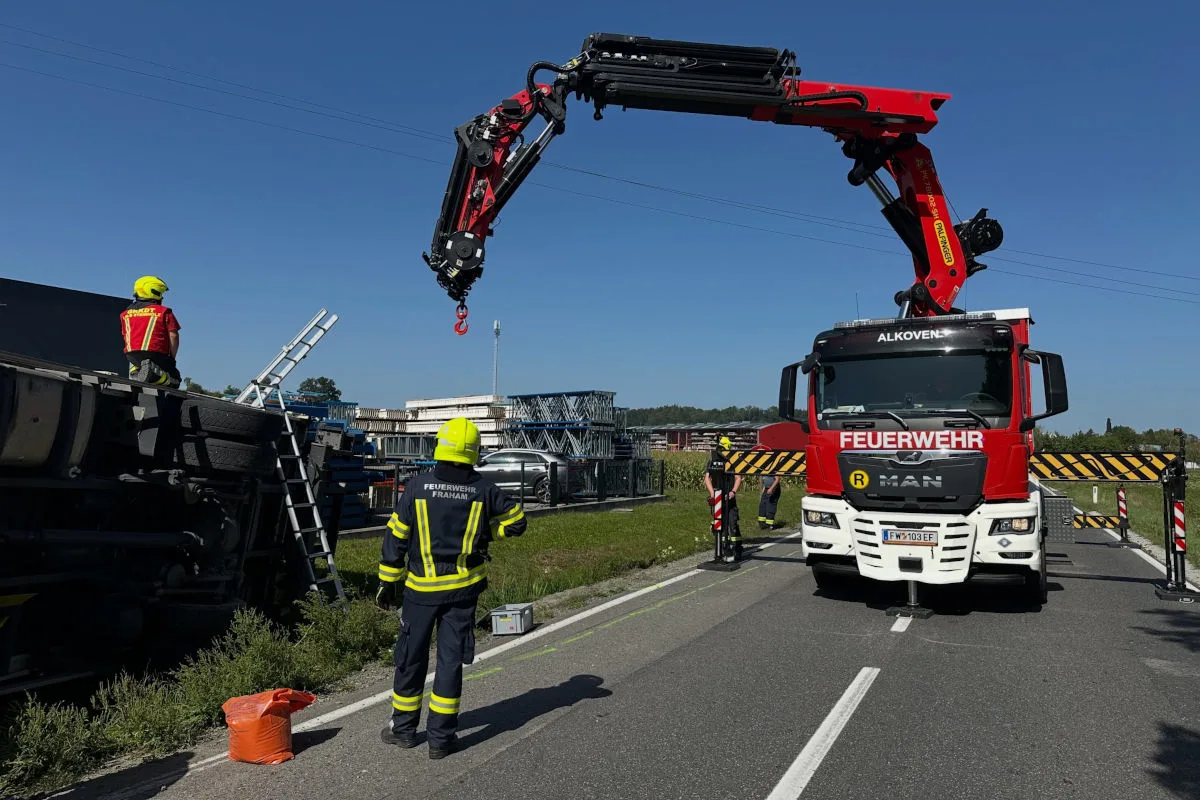 Ein Feuerwehrfahrzeug von Alkoven mit ausgefahrenem Kran steht neben einem umgestürzten Lkw auf einer Straße. Mehrere Feuerwehrleute in Einsatzkleidung sind anwesend.