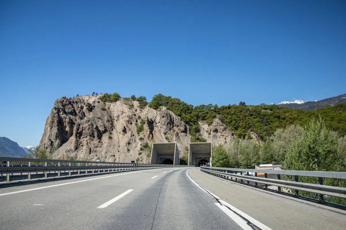 Eine leere Autobahn führt durch zwei Tunnel, die in eine felsige Berglandschaft eingebettet sind. Der Himmel ist klar und blau, deutet auf sonniges Wetter hin.