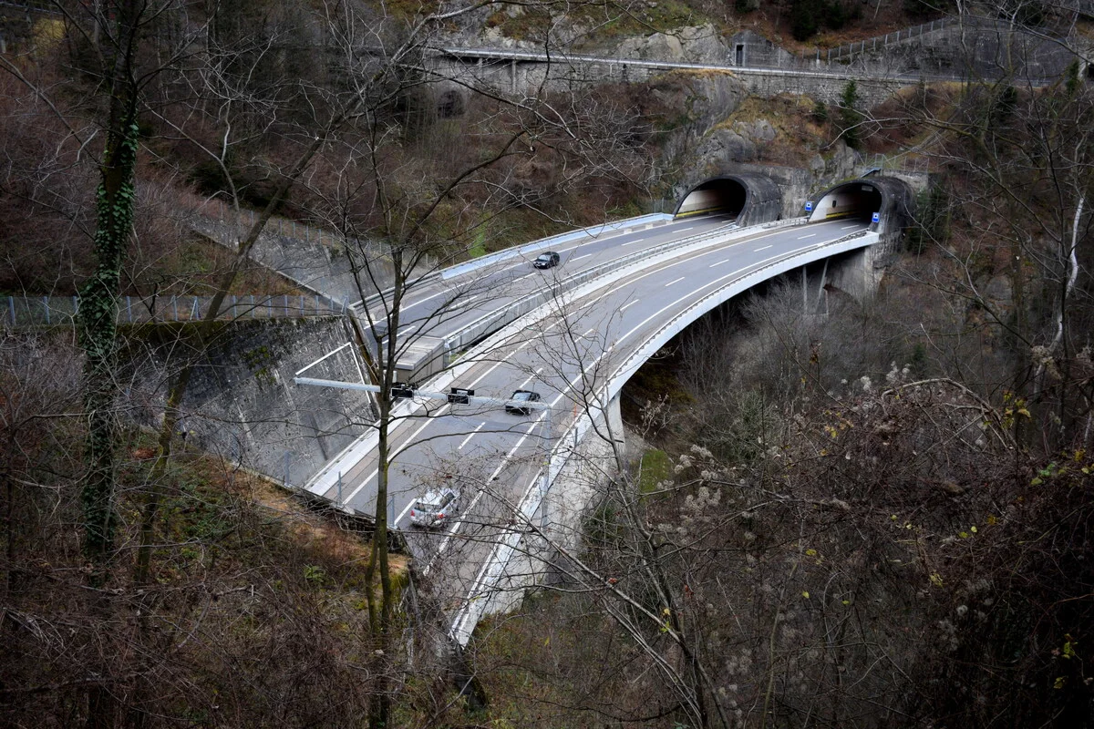 Eine erhöhte Autobahnbrücke führt durch eine bewaldete Schlucht. Mehrere Fahrzeuge sind auf der Brücke zu sehen, und es ist Winter.