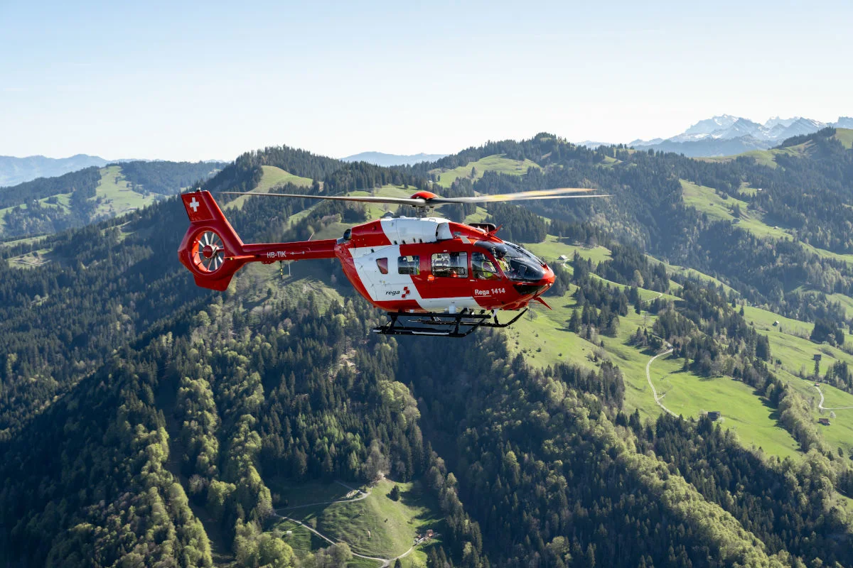 Ein roter Rega-Rettungshelikopter schwebt über einer grünen, hügeligen Landschaft mit bewaldeten Bereichen. Der Himmel ist klar und es ist Tageslicht.