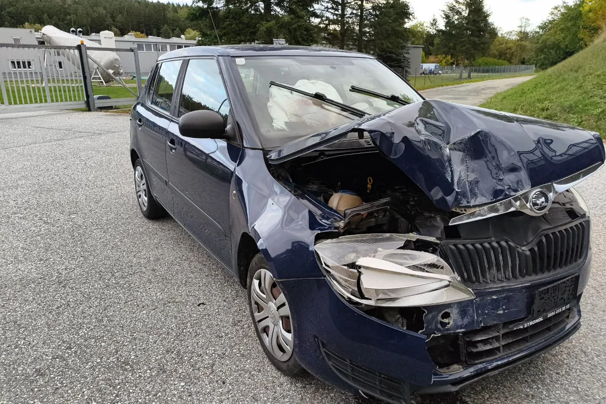 Ein dunkelblaues Auto mit schwer beschädigter Front steht auf einem Parkplatz, im Hintergrund ein Zaun und Industriegebäude.