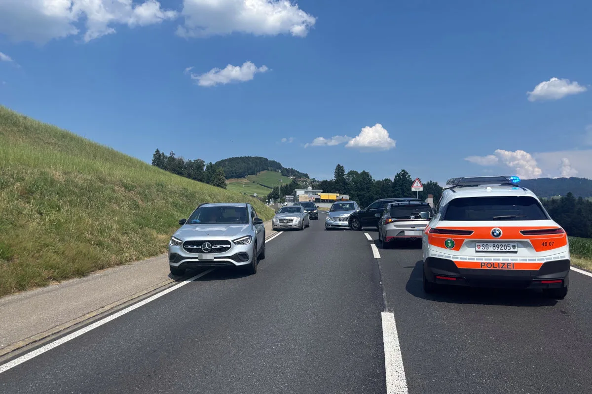 Ein Polizeifahrzeug und mehrere PKWs stehen auf einer Landstraße. Die Szenerie ist ländlich mit Hügeln im Hintergrund.