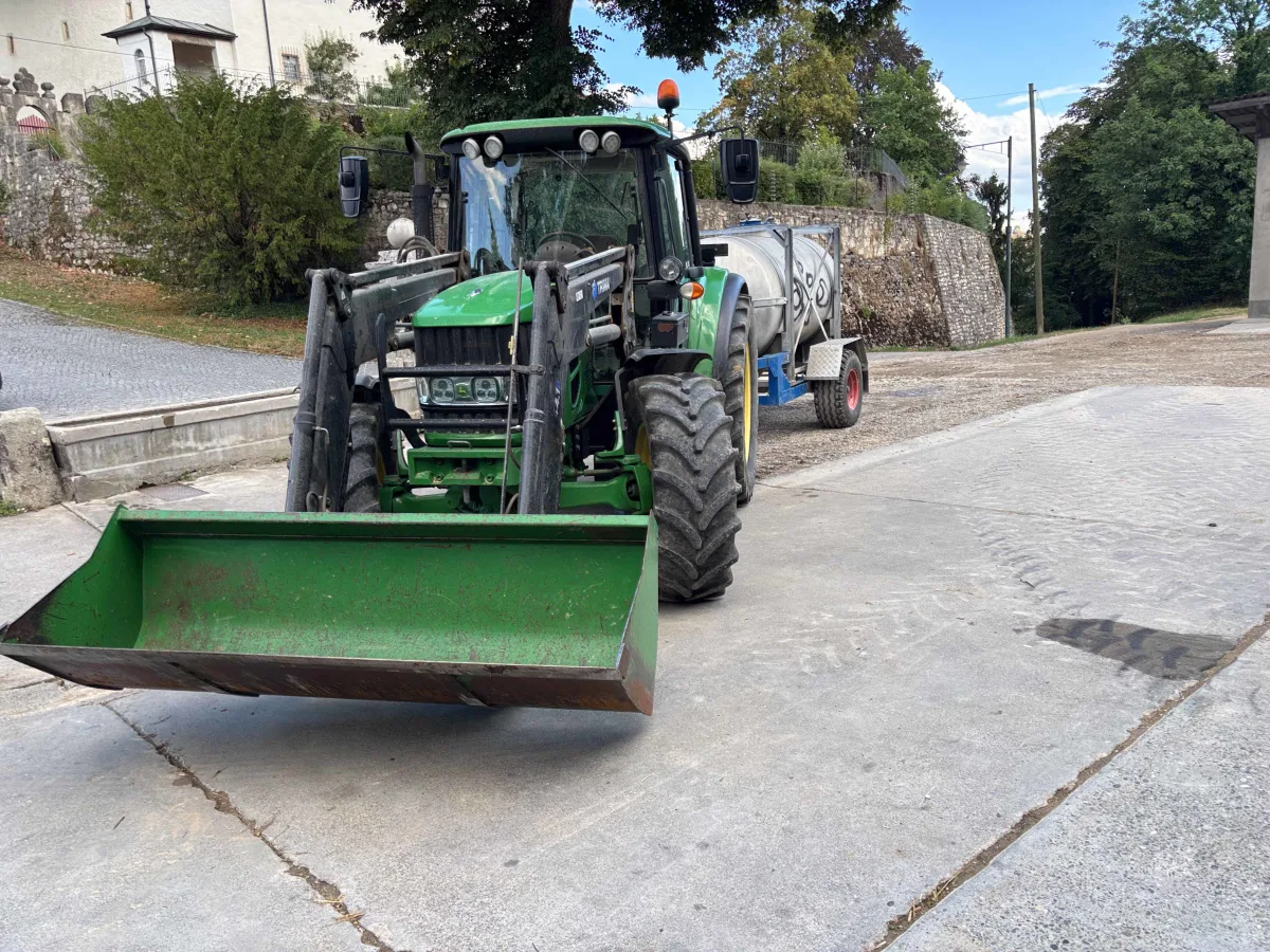 Ein grüner Traktor mit Frontlader steht auf einem gepflasterten Platz in einer ländlichen Umgebung. Bäume und eine Steinmauer sind im Hintergrund sichtbar.