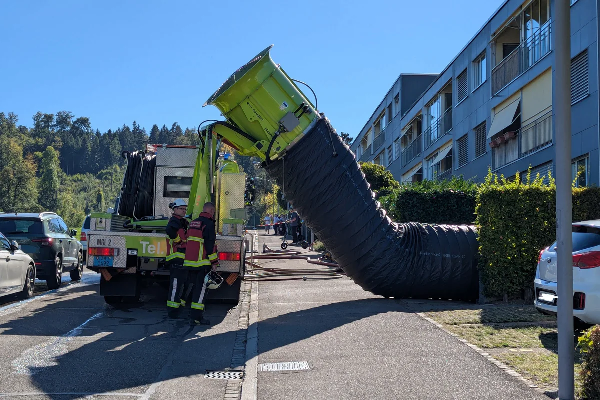 Ein Feuerwehrfahrzeug mit einem angeschlossenen großen schwarzen Lüftungsschlauch steht in einer Wohnstraße. Zwei Feuerwehrleute sind am Fahrzeug aktiv.