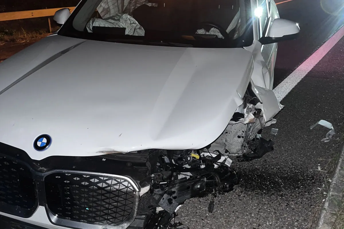 Ein schwer beschädigter weißer BMW steht auf einer Straße. Die Front des Fahrzeugs ist stark eingedrückt, und Teile liegen verstreut auf dem Asphalt.