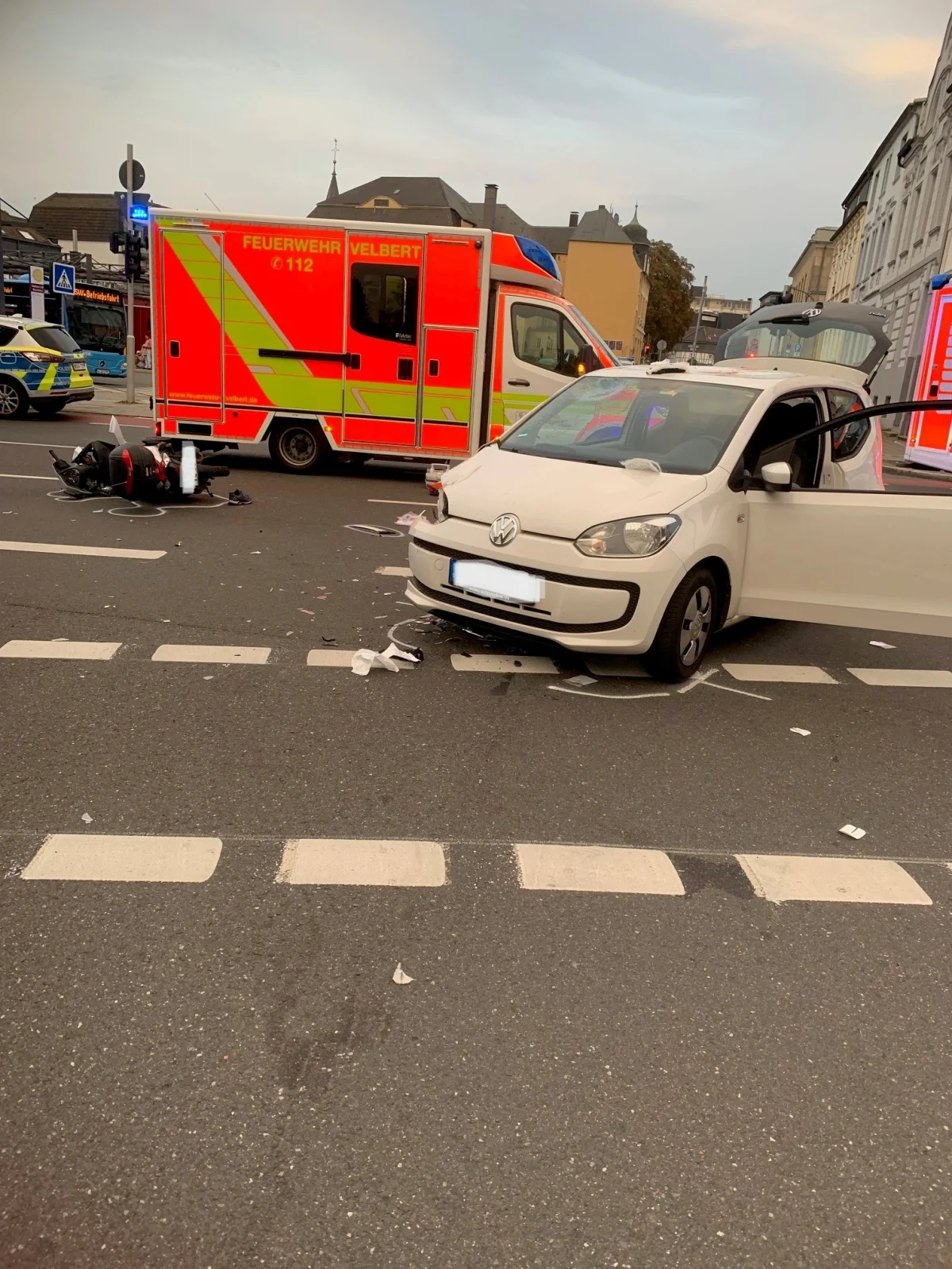 Ein weißer VW steht mit offenem Fahrerfenster auf der Straße, daneben ein gestürztes Motorrad. Im Hintergrund ist ein Rettungsfahrzeug mit der Aufschrift 'FEUERWEHR VELBERT' zu sehen.