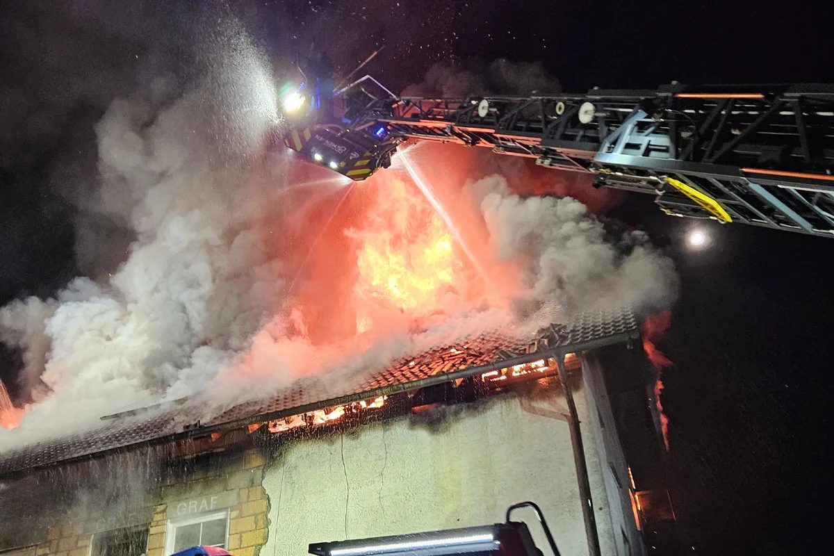 In der Nacht bekämpft die Feuerwehr mit einem wasserführenden Kran einen großen Brand auf einem Dach, während dicker Rauch aufsteigt.