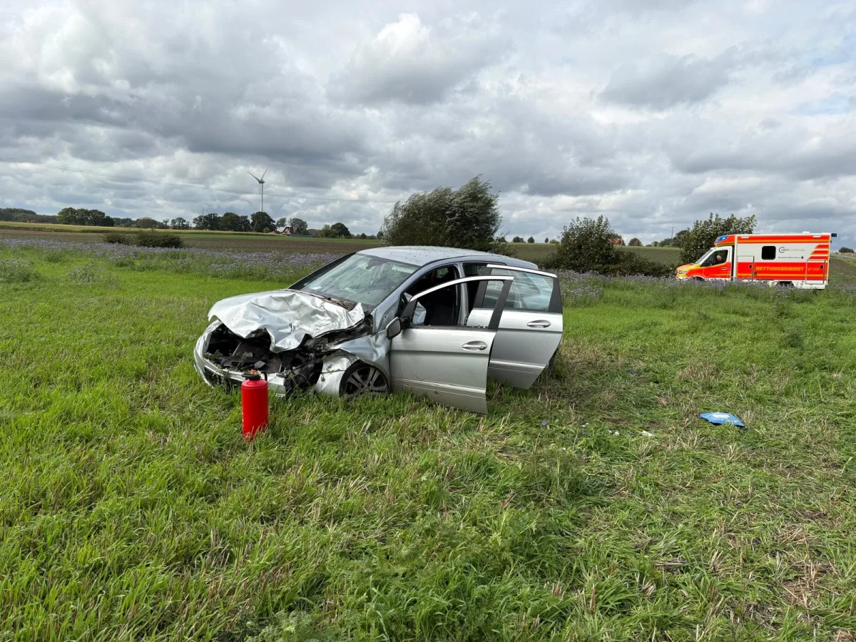 Ein silbernes Auto mit stark beschädigter Front steht auf einer Wiese. Im Hintergrund ist ein Rettungswagen zu sehen.