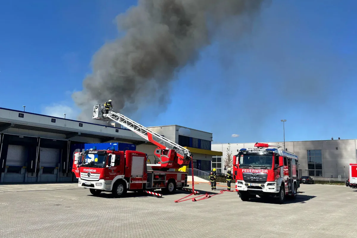 Zwei Feuerwehrfahrzeuge stehen vor einem Gebäude mit starkem Rauch. Eine Drehleiter wird eingesetzt, um den Brand zu bekämpfen.