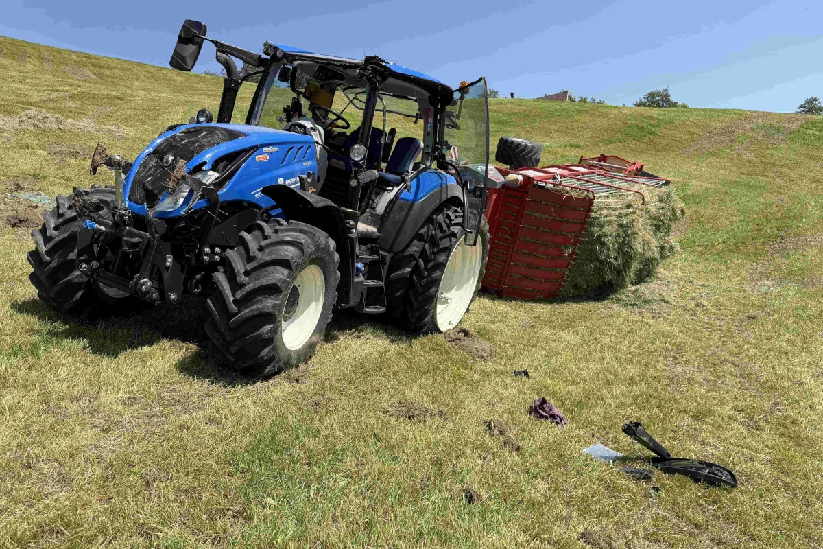 Ein blauer Traktor steht mit geöffneter Fahrertür auf einer Wiese. Hinter ihm liegt eine umgestürzte Ladung Heu.