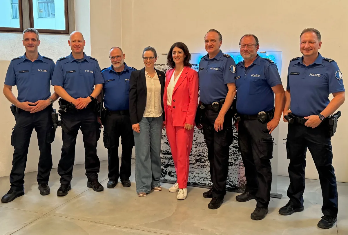 Eine Gruppe von sechs Polizeibeamten in blauen Uniformen posiert neben zwei Frauen. Alle stehen in einem Innenraum.