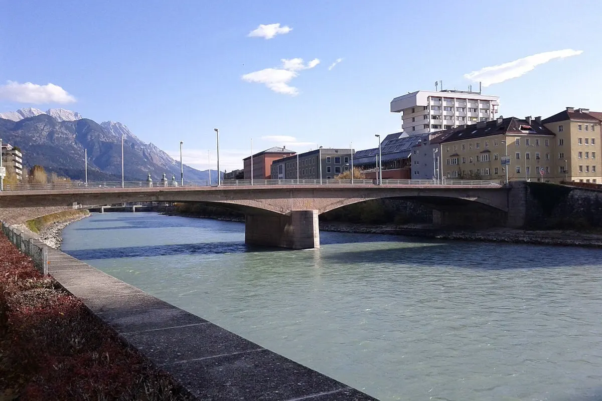 Eine Brücke führt über einen Fluss in Innsbruck, umgeben von Stadtgebäuden und Bergen im Hintergrund. Der Himmel ist klar.