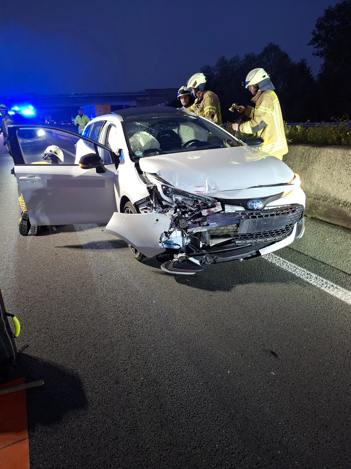 Ein stark beschädigtes weißes Auto steht auf einer Autobahn, umgeben von mehreren Feuerwehrleuten in Uniform.