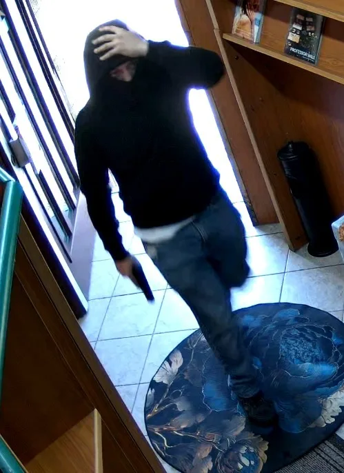 Eine Person mit Kapuze betritt durch eine geöffnete Tür ein Geschäft, während sie einen Gegenstand in der Hand hält. Der Boden ist gefliest und ein runder Teppich ist sichtbar.