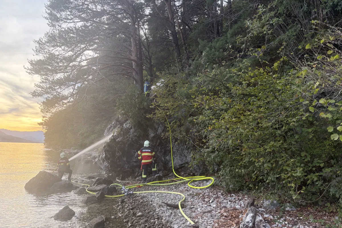 Zwei Feuerwehrleute in Einsatzkleidung bekämpfen mit Schläuchen einen Brand am bewaldeten Ufer eines Sees, während die Sonne untergeht.